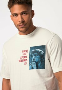 Krémové tričko s krátkým rukávem, na kterém je modrá grafika sochy s červeným textem "SIMPLI - CITY SPEAKS VOLUMES." Měkká bavlněná textilie.