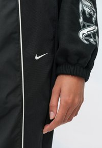 Χέρι ακουμπισμένο στο πλάι, φορούν μαύρο παντελόνι Nike με λευκό λογότυπο swoosh και μαύρο μανίκι με λευκό σχέδιο και ελαστικό μανσέτα.