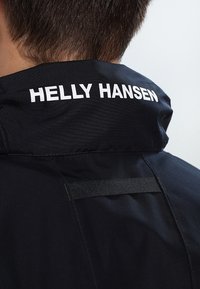 Vue arrière d'une personne portant une veste noire avec le nom de la marque Helly Hansen en blanc sur le col relevé.