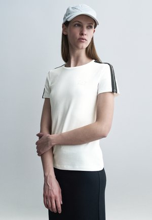 Naine, kes kannab valget t-särki mustade triipudega õlgadel ja valget nokamütsi, seisab ühe käe üle teise rinna peal risti.