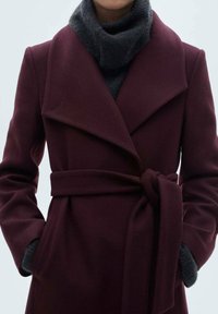 Bordeaux uldfrakke med båndliv, oversize revers og en grå rullekravebluse underneath. Har sidelommer og en struktureret overflade.