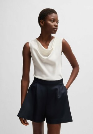Cowl-neck sleeveless top in cream satin paired with high-waisted navy shorts featuring pleats and side pockets. Smooth texture, minimalist design.