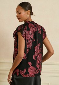 Juoda bliuska su rožiniu gėlių raštu, trumpomis plačiomis rankovėmis, tekstūruota medžiaga ir aukšta apykakle. Paprastas dizainas, laisvas kirpimas, išskirtiniai akcentai nugaroje.