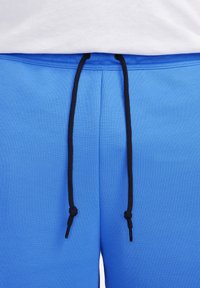 Blå sweatpants med svart dragsko knutna i knutar, bärs med en vit topp synlig vid midjan.