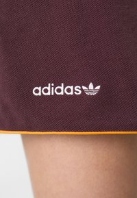 Viininpunaiset urheilushortsit oranssilla reunuksella, joissa on näkyvä valkoinen "adidas"-logo ja trefoil-kuvio, teksturoitua kangasta ja minimalistinen tyyli.