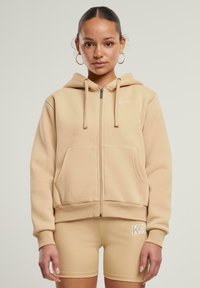 Felpa con zip color beige realizzata in tessuto morbido, con una grande tasca frontale, cappuccio con cordino e logo ricamato sul petto.