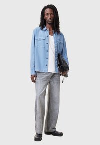 Lichtblauw overhemd met knopen en twee borstzakken, witte t-shirt eronder, grijze brede jeans, zwarte tas en zwarte schoenen.