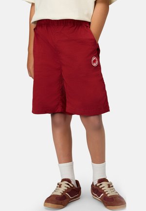 Barn iført røde shorts til knæene med elastisk talje og logo på venstre lår, hvide sokker og brune sneakers med beige snørebånd.