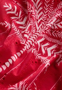 Tissu rouge avec des motifs de fougères blanches, présentant des accents brodés et des découpes. Texture lisse, drapée en plis, mettant en valeur un design complexe.