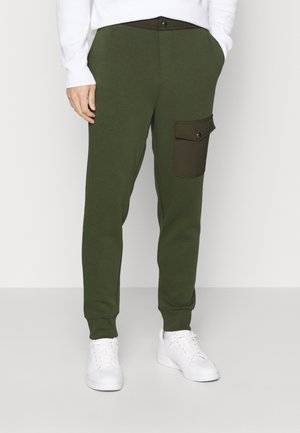Homme portant un pantalon de jogging vert olive avec une poche plaquée sombre sur la cuisse droite et des baskets blanches.