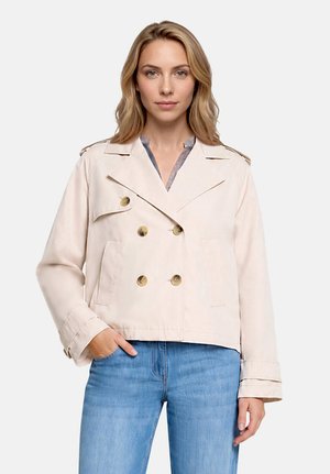 Femme portant une veste beige à double boutonnage avec des boutons dorés et un jean bleu, debout devant un fond uni clair.