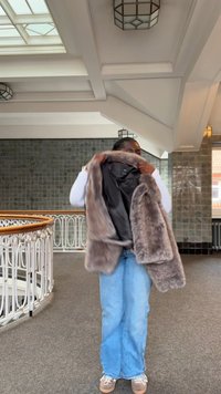 Kamizelka z futerka sztucznego w jasnobrązowym kolorze z czarną podszewką, noszona na białej bluzce i niebieskich dżinsach. Teksturowany, miękki zewnętrzny materiał z otwartym przodem.