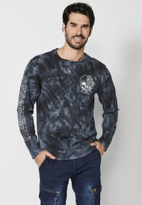 Hosszú ujjú póló sötét tie-dye mintázatban, pamutból készült. A mellkason és a bal ujján koponya grafika és felirat található.