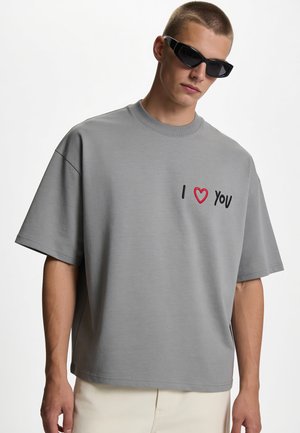 Jeune homme portant des lunettes de soleil noires et un t-shirt gris oversize avec le texte "I heart you" sur la poitrine, debout devant un fond uni.