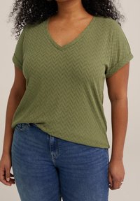 Grön kortärmad topp med V-halsringning och fiskbensmönster, kombinerad med blå jeans. Avslappnad passform, textil med struktur.