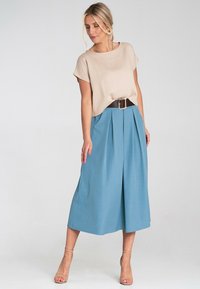 Beżowy krótki top i plisowana, średniej długości niebieska spódnica z czarnym paskiem. Gładka tekstura, lekki materiał i swobodny fason. Czyste tło.