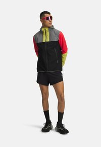 Mønstret jakke i grå, rød, sort og neongrønne sektioner, med lynlås-lommer. Bæres med sorte shorts og sportsko.