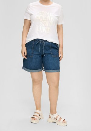 Person trägt ein weißes kurzärmeliges Shirt mit Text, blaue Denim-Shorts mit Kordelzug und weiße klobige Sandalen und steht vor einem einfarbigen Hintergrund.