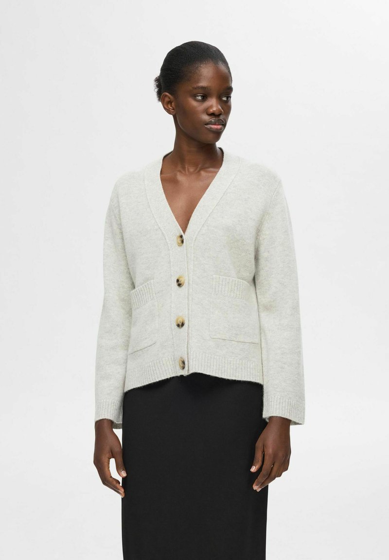 Šviesiai pilkas megztinis su V formos iškirpte, raibomis rankogaliais ir dviem priekinėmis kišenėmis. Priekyje yra dideli rudi mygtukai.