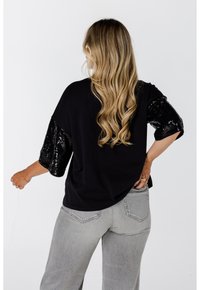 Zwarte top met pailletten mouwen, relaxed fit en afgeronde onderkant, gecombineerd met lichtgrijze jeans. De textuurcontrasten tussen matte en glanzende oppervlakken.