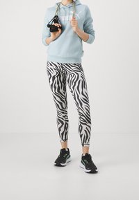 Felpa leggera blu con logo bianco, leggings con stampa zebra bianca e nera e scarpe sportive nere con accenti verdi.