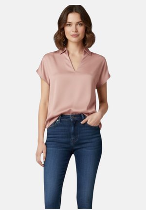 Žena nosí svetloružovú blúzku s krátkym rukávom, zastrčenú do tmavomodrých džínov s vysokým pásom, stojí s jednou rukou v vrecku.