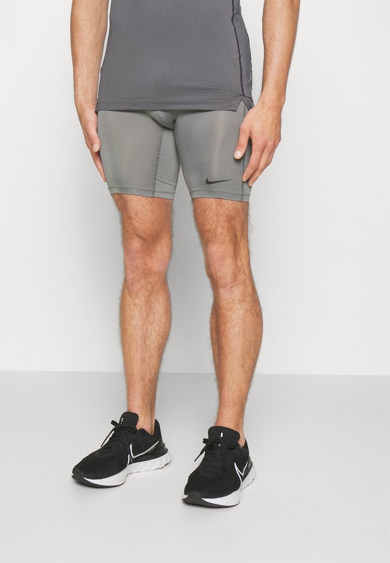 Homme portant un short de compression gris Nike et des chaussures de running noires Nike, debout devant un fond uni.