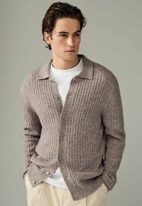 Cardigan bej deschis cu nervuri, fabricat din lân�ă moale, prezentând un sistem de închidere cu nasturi, un guler clasic și o croială lejeră peste o cămașă albă.