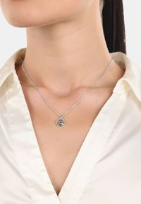 Collana d'argento con un pendente circolare che presenta una gemma centrale, circondata da un anello di piccole pietre, indossata sopra una camicia di colore chiaro.