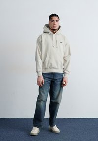 Felpa leggera color beige con tasca frontale e coulisse, abbinata a jeans blu sbiaditi e sneakers color crema. Look semplice e vestibilità rilassata nel complesso.