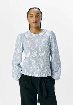 Himmelbluse mit langen Ärmeln und weißen Blumenmustern, runder Halslinie und gerafften Ärmeln. Kombiniert mit schwarzen Hosen.
