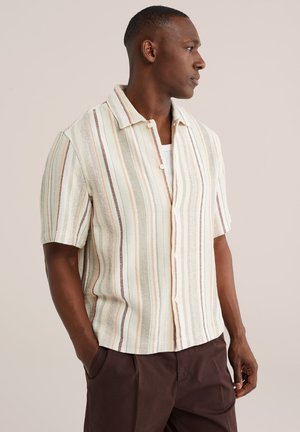 Mand iført kortærmet, beige og brunstribet skjorte med knapper og brune bukser, stående med hænderne i lommerne, vendt mod højre.