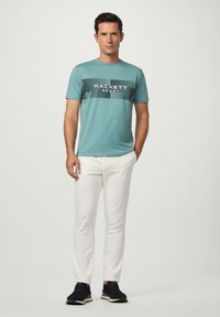 Muž oblečený v tyrkysovém sportovním tričku Hackett, bílých kalhotách a černých teniskách stojící proti hladkému světlému pozadí.