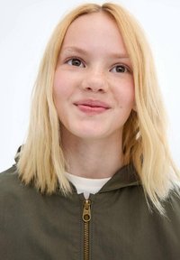 Olivově zelená zipová bunda s kapucí, vybavená zlatým zipem a lehkým, hladkým povrchem. Modelka má rovné, světle blond vlasy.