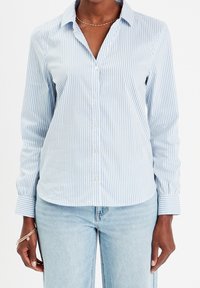 Femme portant une chemise à boutons à rayures verticales bleu clair et blanches avec manches longues et un jean bleu clair, accessoirisée de bracelets et d'un collier.