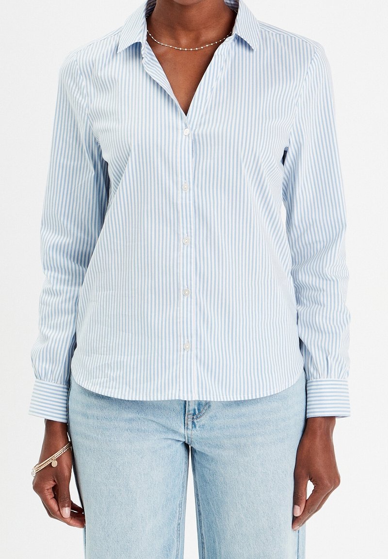 Femme portant une chemise à boutons à rayures verticales bleu clair et blanches avec manches longues et un jean bleu clair, accessoirisée de bracelets et d'un collier.