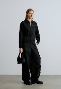 Felpa nera con zip e collo alto, abbinata a pantaloni cargo neri larghi. La modella tiene una piccola borsa nera e indossa scarpe nere.