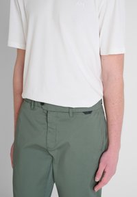 T-shirt in cotone bianco leggero abbinato a pantaloni in cotone verde scuro. I pantaloni presentano un fronte piatto con chiusura a bottone e un dettaglio di branding sottile.