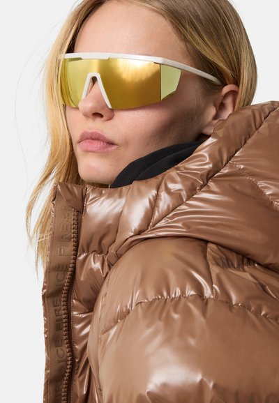 Kvinde med langt blond hår iført reflekterende guld-solbriller og en skinnende brun puffjakke med høj krave mod en hvid baggrund.