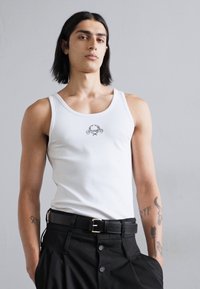 Valge ribastatud tank top ringja kaelusega ja väikese graafika kujundusega rinnal, koos kõrge vöökohtadega mustade pükstega ja vööga.
