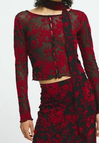 Haut en dentelle fleurie rouge à manches longues, avec un devant à boutons, un ourlet cropé, porté avec une jupe assortie présentant un motif floral similaire.