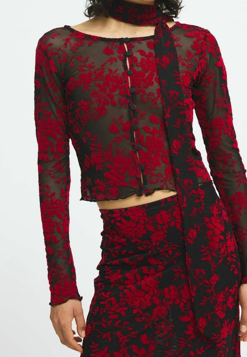 Haut en dentelle fleurie rouge à manches longues, avec un devant à boutons, un ourlet cropé, porté avec une jupe assortie présentant un motif floral similaire.