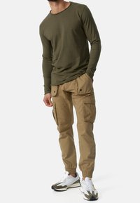 Lange mouwen olijfgroen overhemd gecombineerd met beige cargobroek met zijzakken en elastische manchetten, aangevuld met witte en groene sneakers.