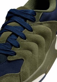 Αθλητικό παπούτσι από βελούδο σε ελιά πράσινο και ναυτικό μπλε με σκουρόμπλε κορδόνια, υφή με στρώσεις στο πάνω μέρος και  λεπτομέρειες λευκής καουτσούκ σόλας.