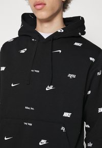 Sudadera negra con logos blancos de Nike dispersos, con bolsillo tipo canguro y capucha con cordón. Textura suave y diseño casual.