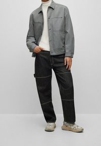 Graue Hemdjacke über einem weißen Rollkragenpullover, kombiniert mit dunklen Jeans mit sichtbaren Nähten. Beigefarbene, strukturierte Sneakers vervollständigen das Outfit.