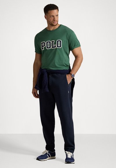 T-shirt de algodão verde com "POLO" em letras azul-marinho; calças de moletão azul-marinho; suéter de malha azul-marinho amarrado à cintura; sapatos de desporto azuis com atacadores brancos.