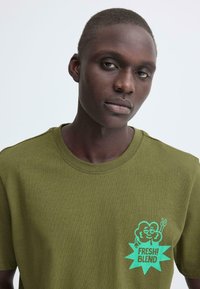 Olijfgroene katoenen t-shirt met een gestructureerde afwerking en een turquoise afbeelding met de tekst "FRESH! BLEND" en een cartoonwolkje.