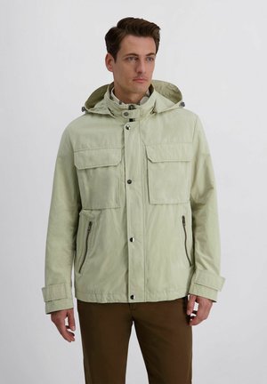 Man draagt een lichtbeige capuchonjas met drukknoopsluiting aan de voorkant en ritssluitingen op de zakken, gecombineerd met een bruine broek, staand tegen een effen achtergrond.