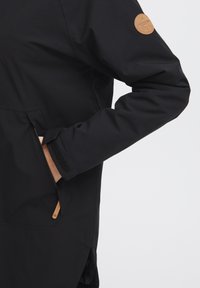 Black jacket with a smooth texture, featuring a zippered pocket and a brown leather logo patch on the sleeve.